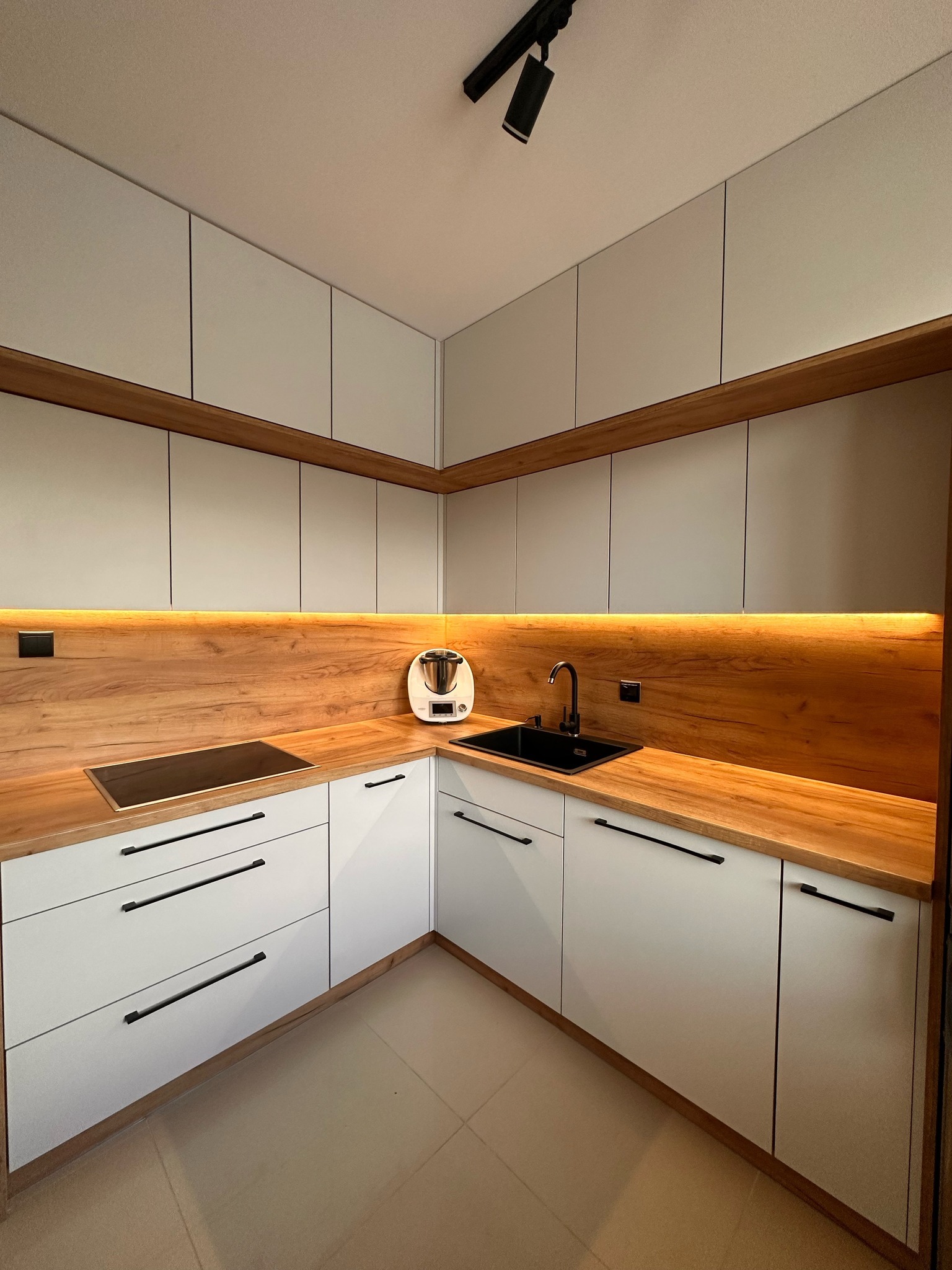 Nowoczesna kuchnia narożna z białymi szafkami, drewnianym blatem z podświetleniem LED, czarną armaturą i płytą indukcyjną.