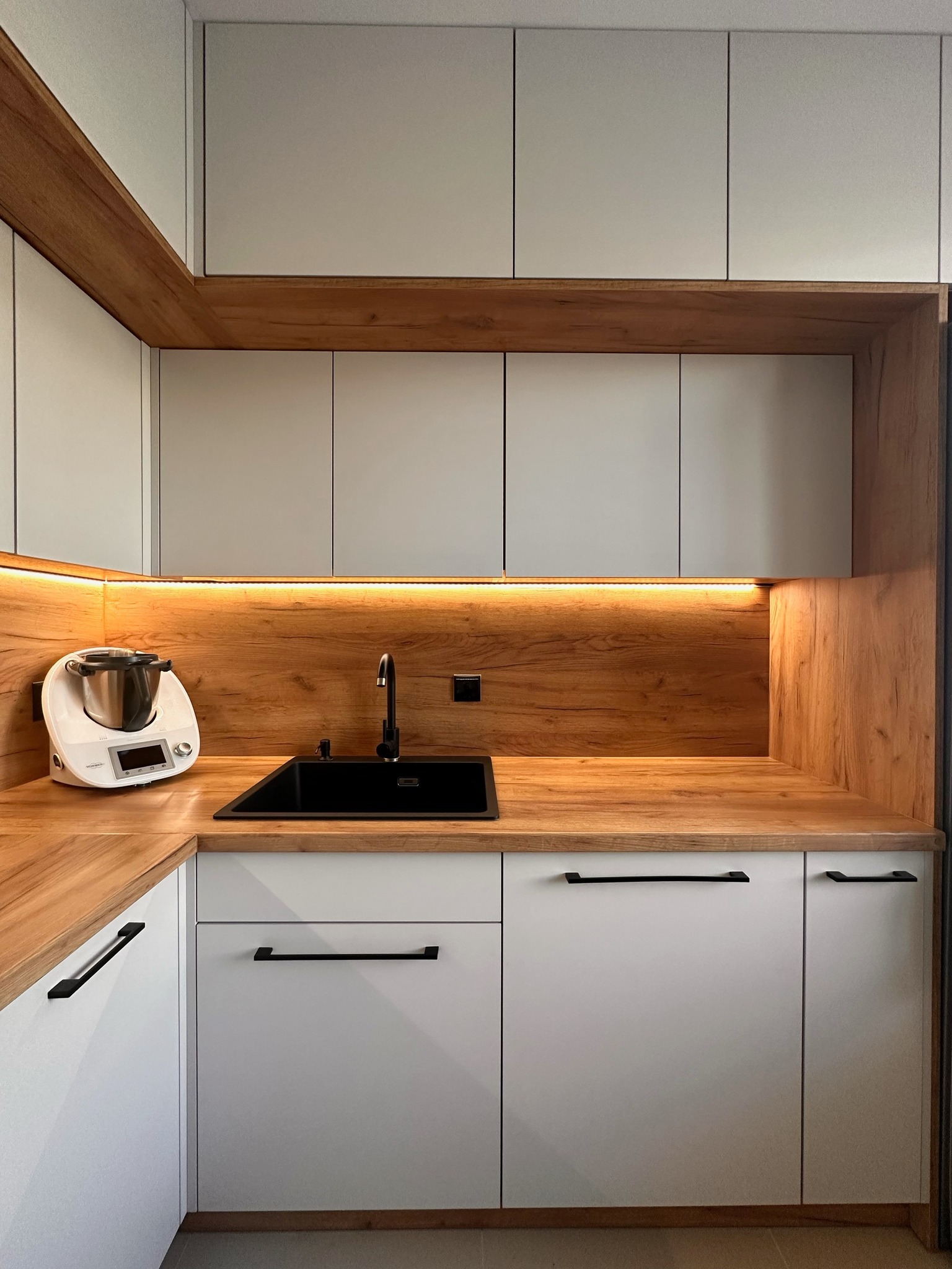 Nowoczesna kuchnia z białymi frontami szafek, drewnianym blatem i panelem ściennym, podświetleniem LED oraz czarną armaturą i zlewozmywakiem, z widocznym robotem kuchennym.