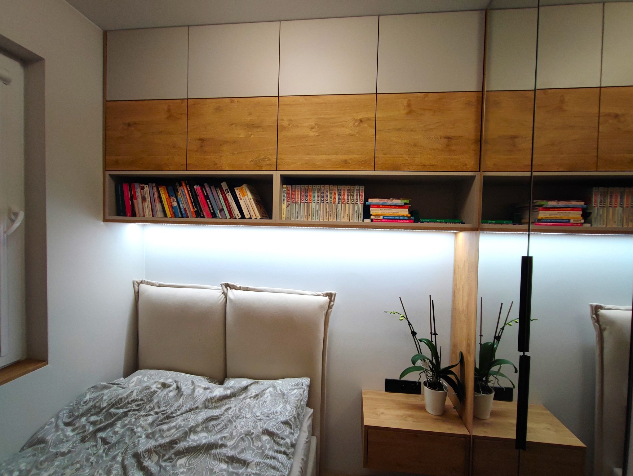 Sypialnia z zabudową meblową: szafki wiszące z drewnianymi frontami i otwartą półką z książkami, podświetlenie LED, łóżko z tapicerowanym zagłówkiem, stolik nocny z orchideami.
