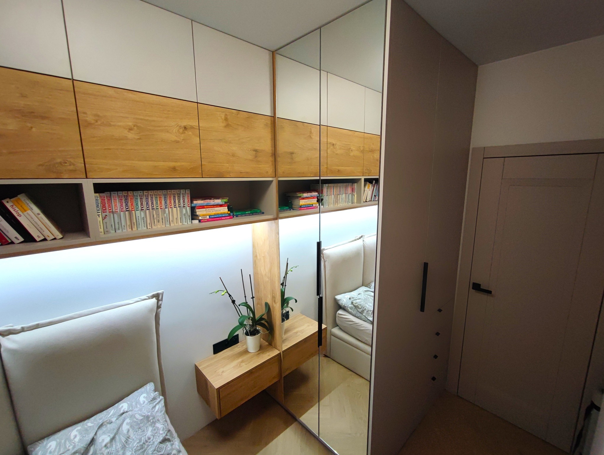 Nowoczesna szafa wnękowa z lustrzanymi drzwiami przesuwnymi i półkami na książki, oświetlenie LED podkreślające drewniane i białe fronty, szafka nocna z kwiatem, odbicie łóżka z szarym zagłówkiem.