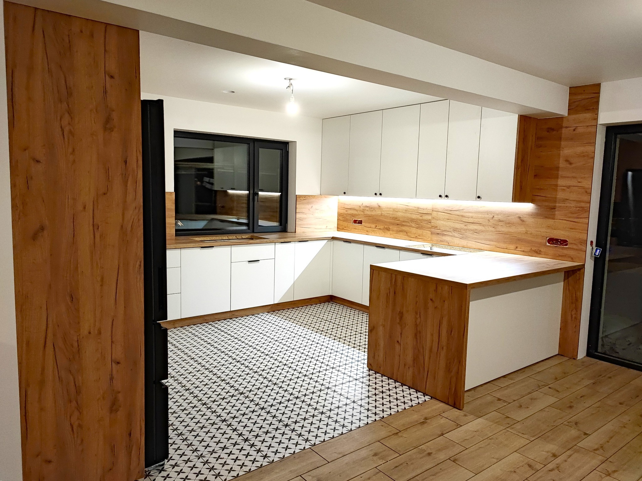 Nowoczesna kuchnia z białymi szafkami, drewnianymi akcentami i wzorzystą podłogą, widziana z perspektywy wejścia.