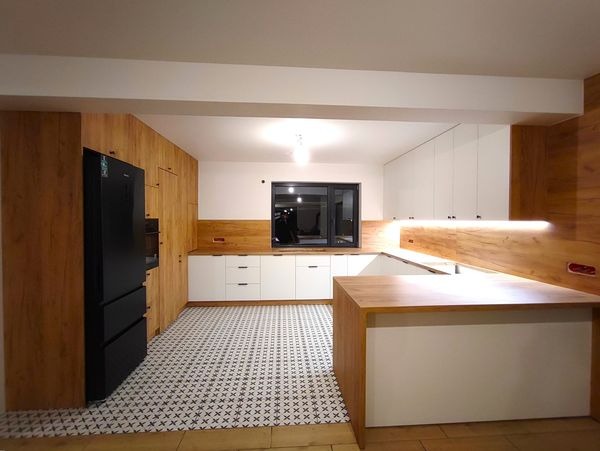 Nowoczesna kuchnia z białymi szafkami, drewnianymi akcentami i wzorzystą podłogą, oświetlona listwą LED pod blatem.