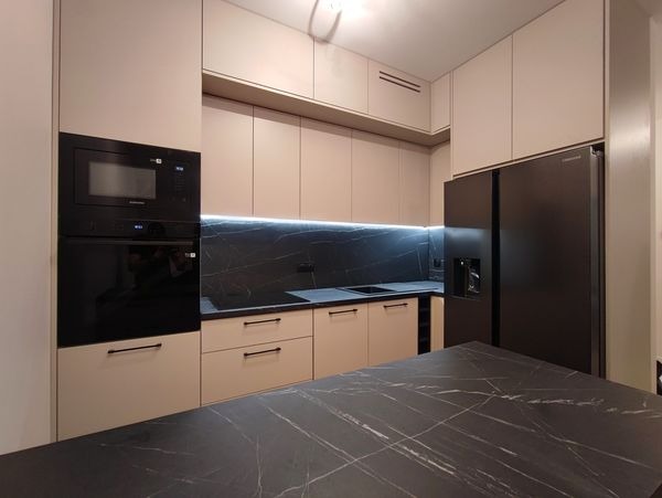 Nowoczesna kuchnia z beżowymi szafkami, czarnym blatem z marmurowym wzorem i wbudowanym sprzętem AGD, takim jak piekarnik i lodówka.