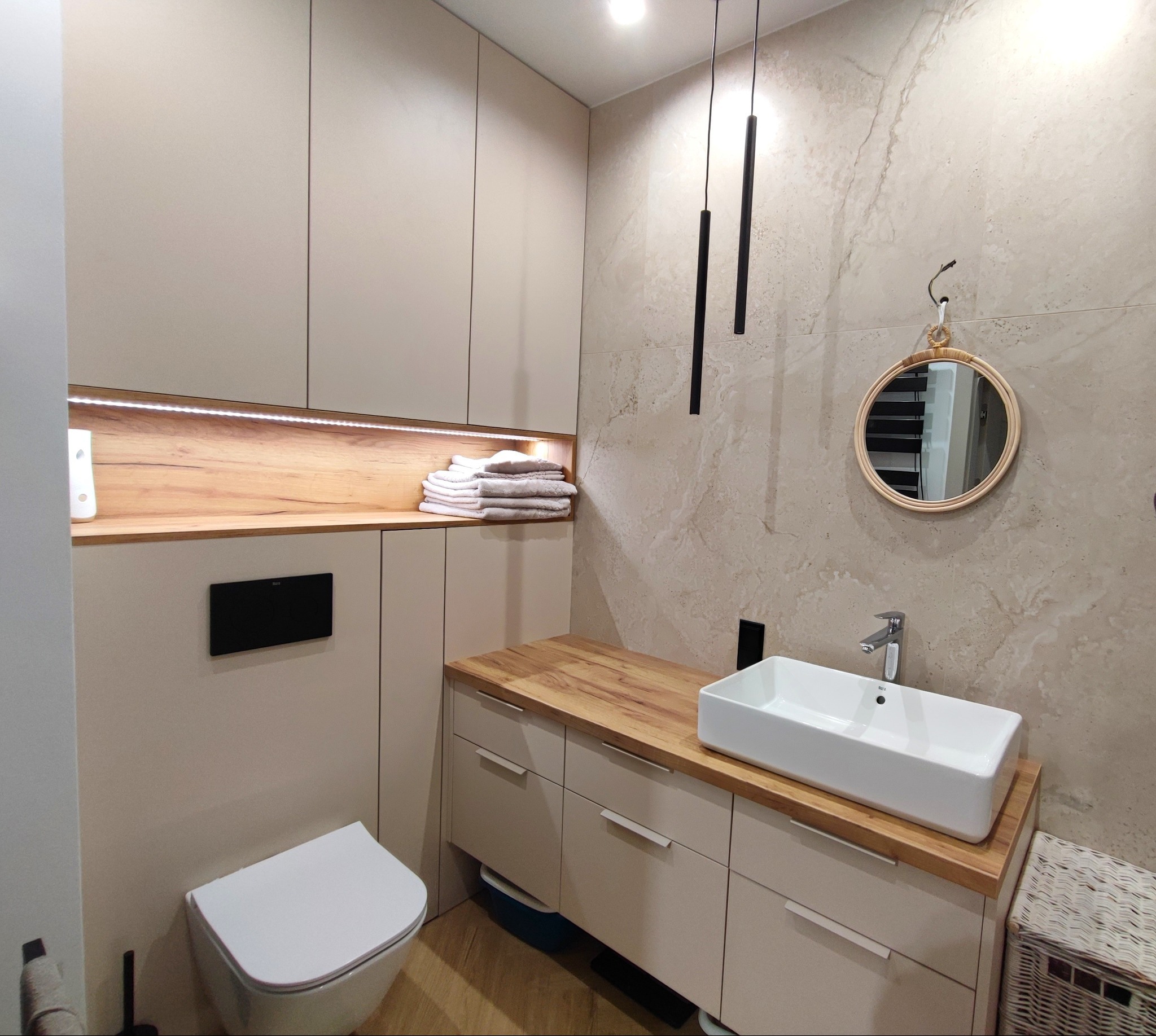 Nowoczesna łazienka z beżowymi szafkami, drewnianym blatem, białą umywalką i okrągłym lustrem w ramie z rattanu, oświetlona wiszącymi lampami tubowymi.