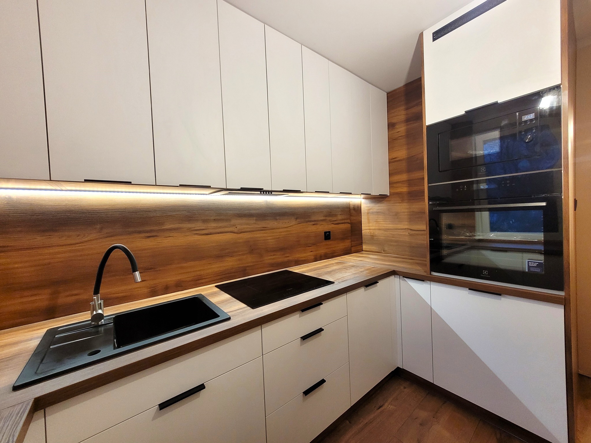 Nowoczesna kuchnia z białymi frontami szafek, drewnianym blatem i podświetleniem LED, czarny zlew z elastyczną wylewką, piekarnik i mikrofalówka w zabudowie.