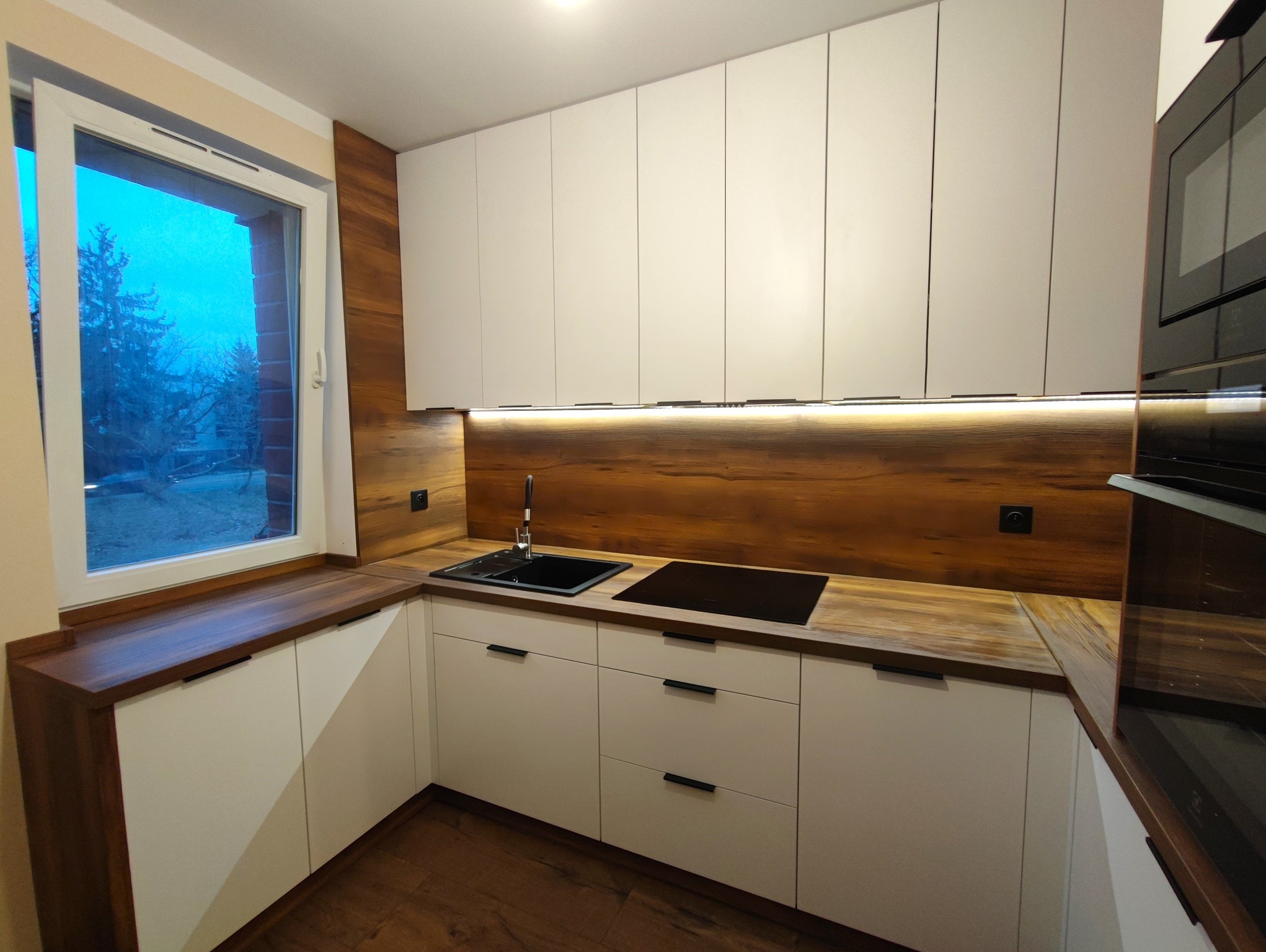 Nowoczesna kuchnia w kształcie litery L z białymi frontami szafek, drewnianym blatem i panelem nad blatem, czarnym zlewem i płytą indukcyjną, oświetleniem LED pod szafkami wiszącymi, z widokiem...