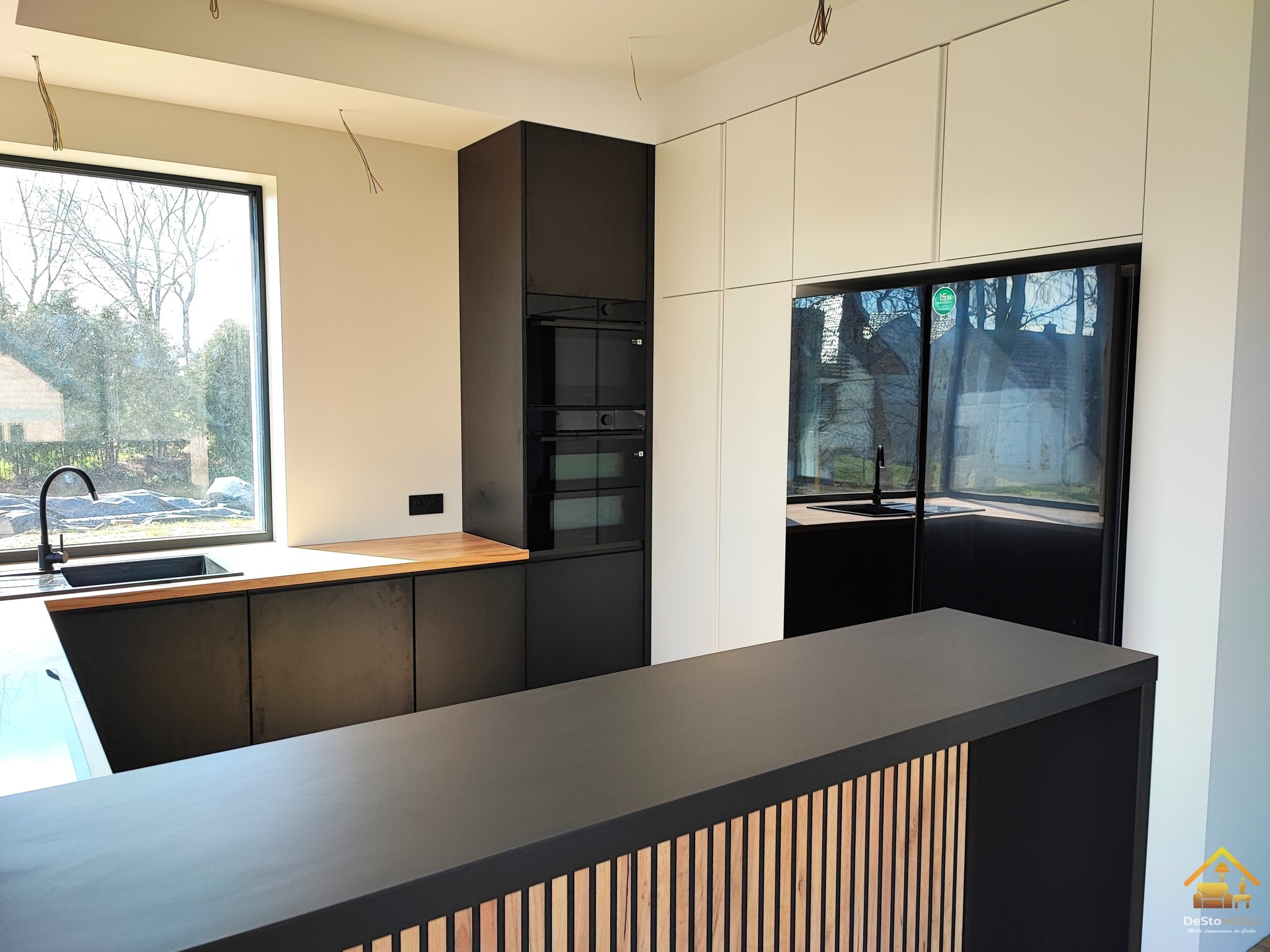 Nowoczesna kuchnia z czarnymi szafkami dolnymi i białymi górnymi, drewniany blat i wyspa z dekoracyjnymi pionowymi listwami, widok na okno z zielenią na zewnątrz, w trakcie montażu - widoczne kable.
