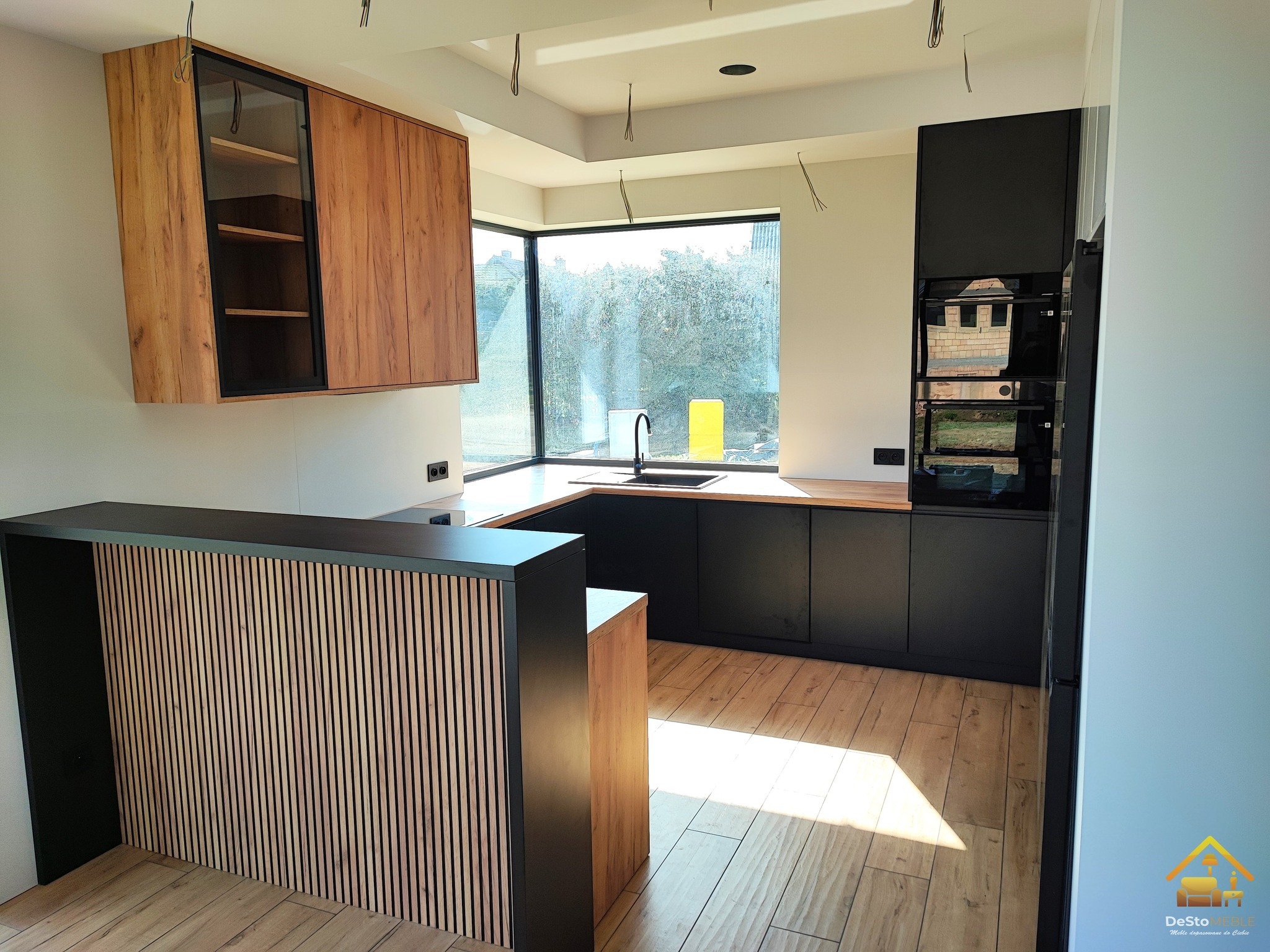 Nowoczesna kuchnia z czarnymi matowymi szafkami dolnymi i drewnianymi górnymi, wyspa z lamelami, jasna podłoga, duże okno z widokiem na zieleń, minimalizm.