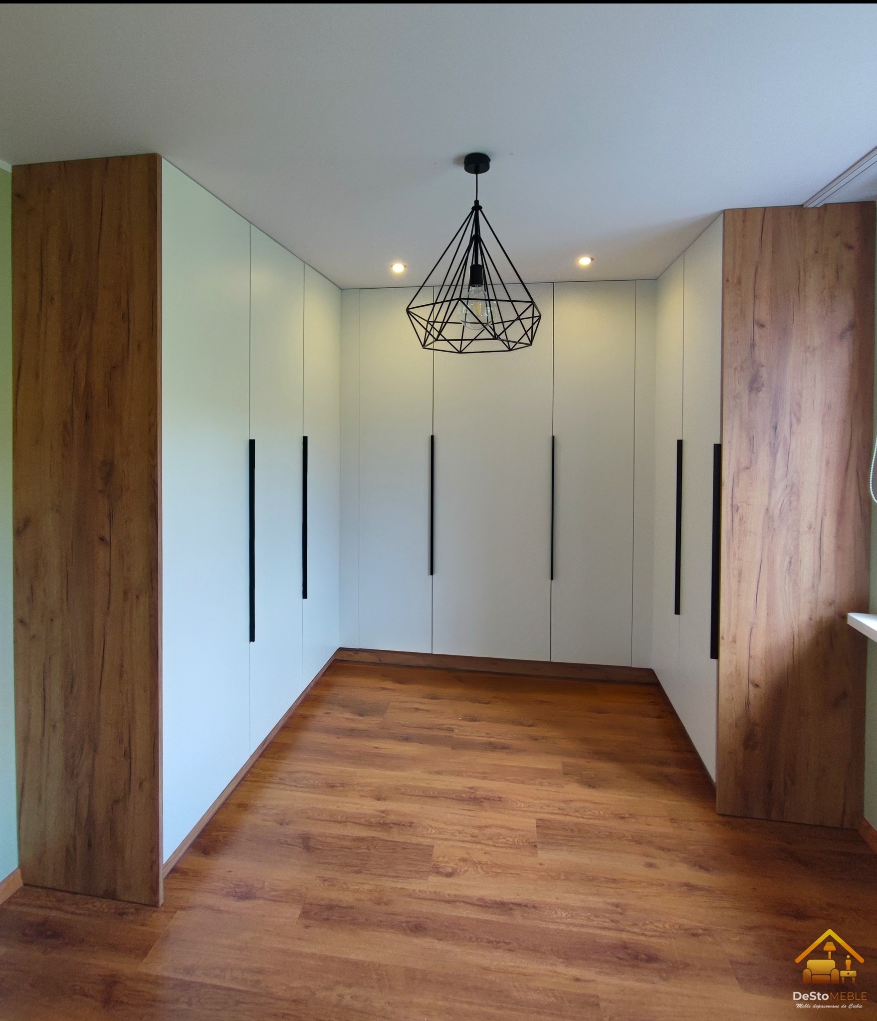 Przestronna garderoba z białymi frontami i drewnianymi bokami, czarne, długie uchwyty, brązowa podłoga, nowoczesna lampa wisząca z geometrycznym kloszem.