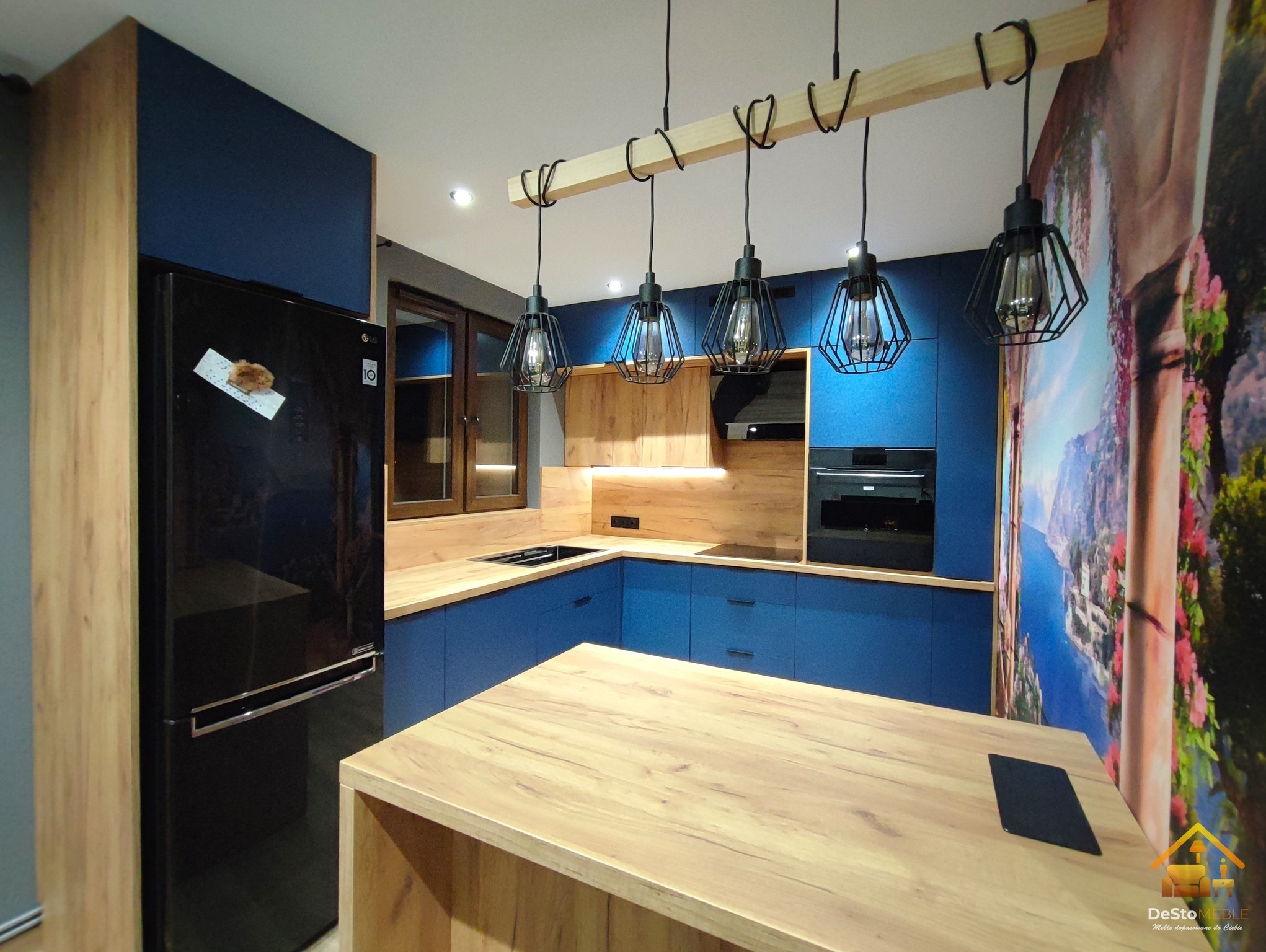 Nowoczesna kuchnia z niebieskimi frontami szafek, drewnianymi blatami i wyspą, oświetlona lampami w metalowych kloszach, z lodówką obok szafy w kolorze drewna i fototapetą z motywem krajobrazowym.