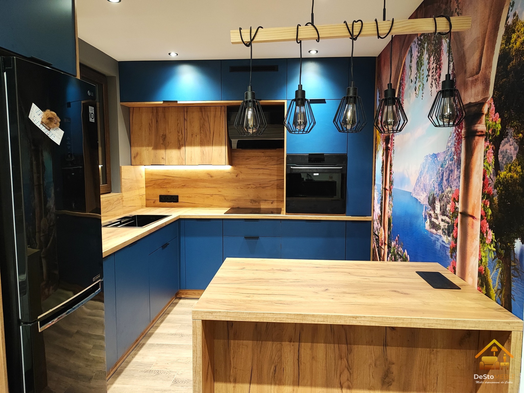 Nowoczesna kuchnia z niebieskimi frontami szafek i drewnianymi blatami, wyspą kuchenną, czarnym piekarnikiem i lodówką, oświetlona trzema wiszącymi lampami w stylu industrialnym oraz z fototapetą...