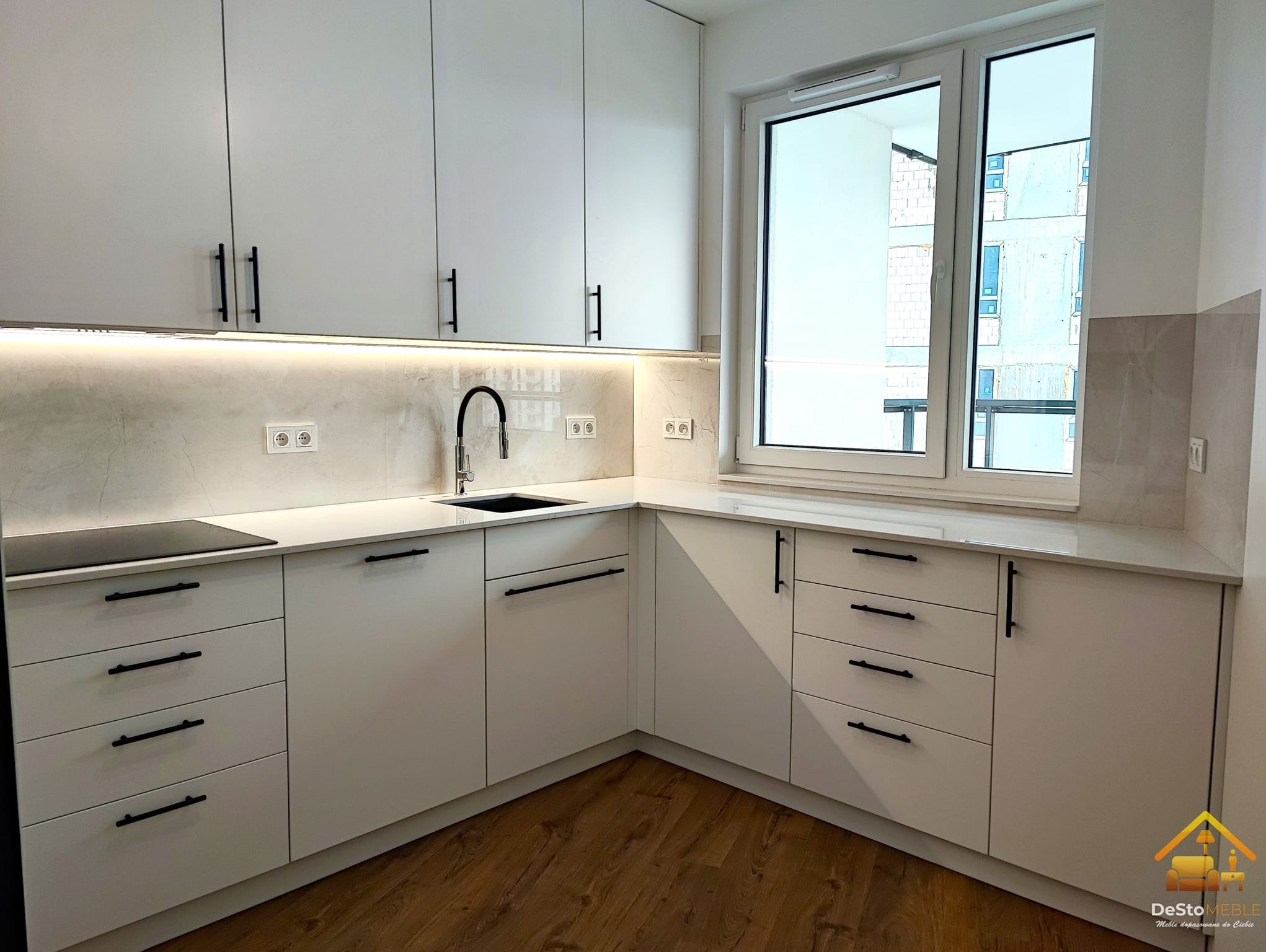 Nowoczesna, jasna kuchnia z białymi frontami, czarnymi uchwytami i blatem imitującym marmur, oświetlenie LED podszafkowe, widok na budynek w trakcie budowy przez okno.