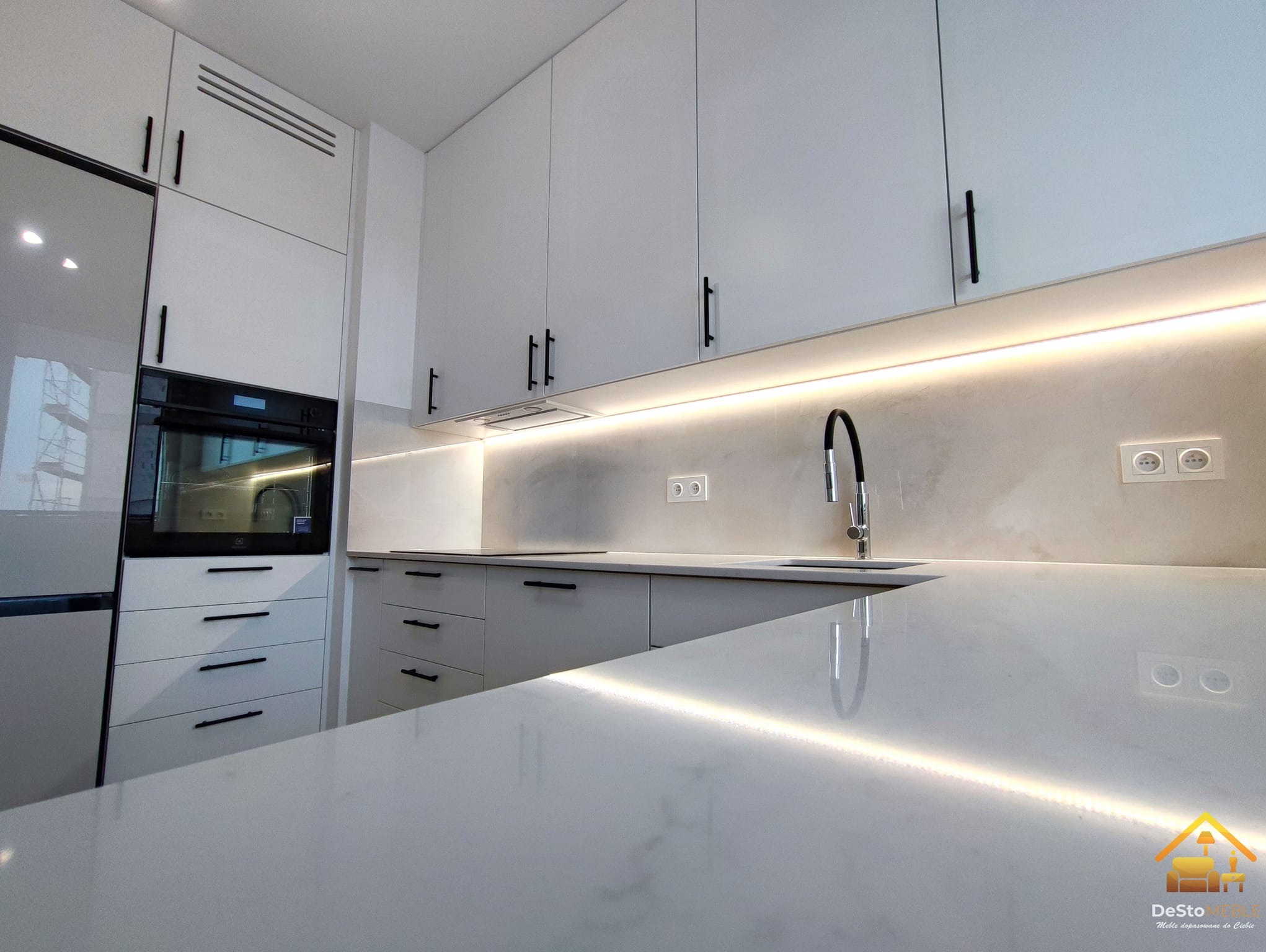 Minimalistyczna, biała zabudowa kuchenna z czarnymi uchwytami, podświetlana LED listwą podszafkową, widok na blat z połyskiem i zlew z czarną baterią.