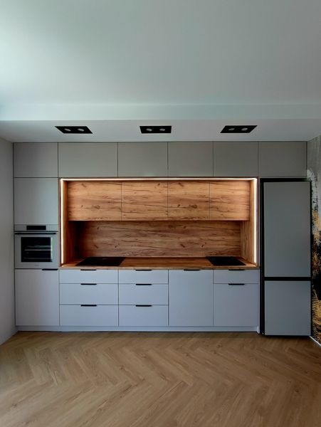 Nowoczesna kuchnia z szarymi frontami i drewnianym panelem nad blatem roboczym, oświetlona listwami LED, z parkietem ułożonym w jodełkę.