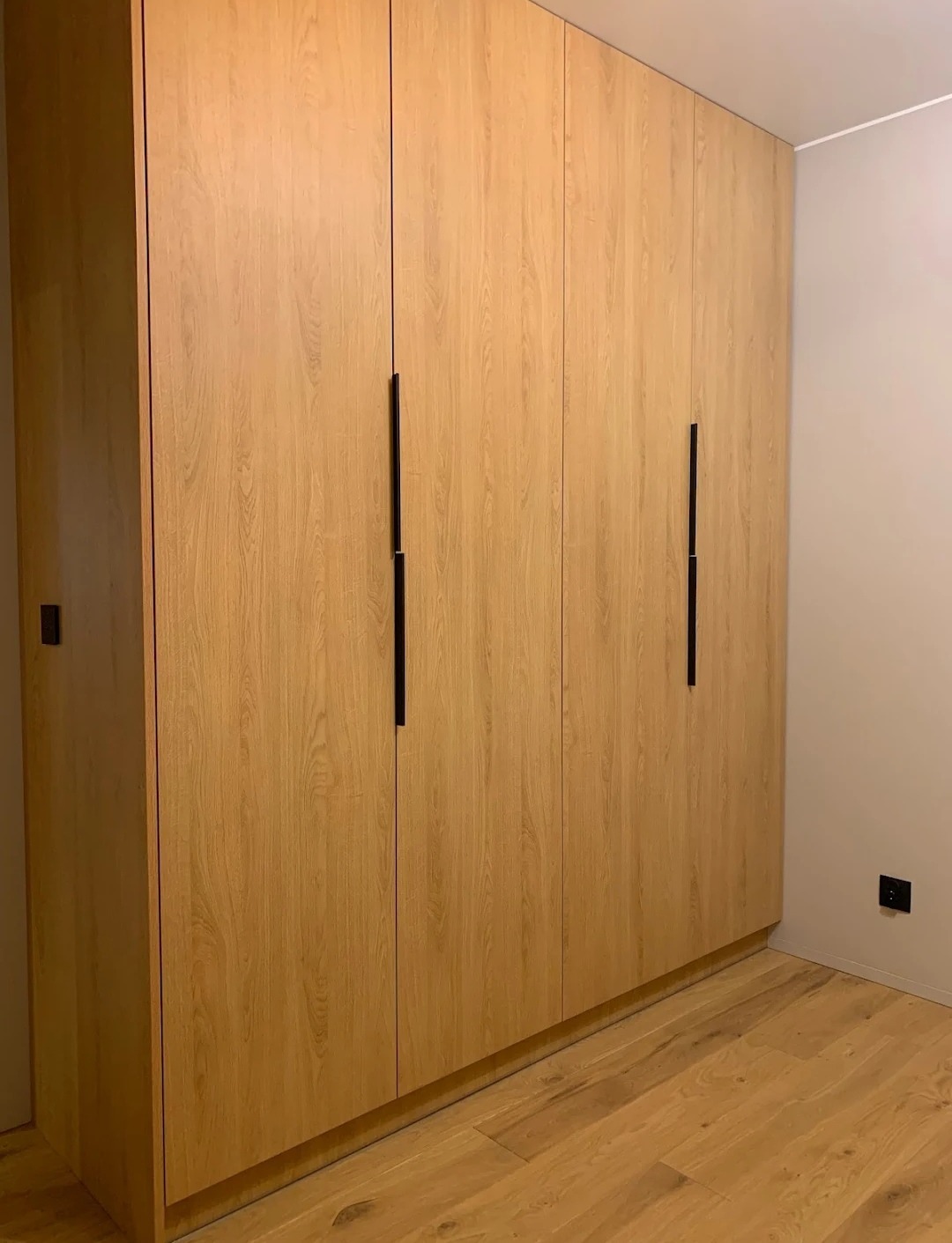 Drewniana szafa wnękowa z czterema drzwiami i minimalistycznymi, czarnymi uchwytami, dopasowana kolorystycznie do drewnianej podłogi, widoczny kontakt elektryczny w rogu pomieszczenia.