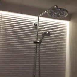 Chromowana bateria prysznicowa z deszczownicą i słuchawką na tle ściany z poziomymi, falowanymi płytkami podświetlonymi od góry.