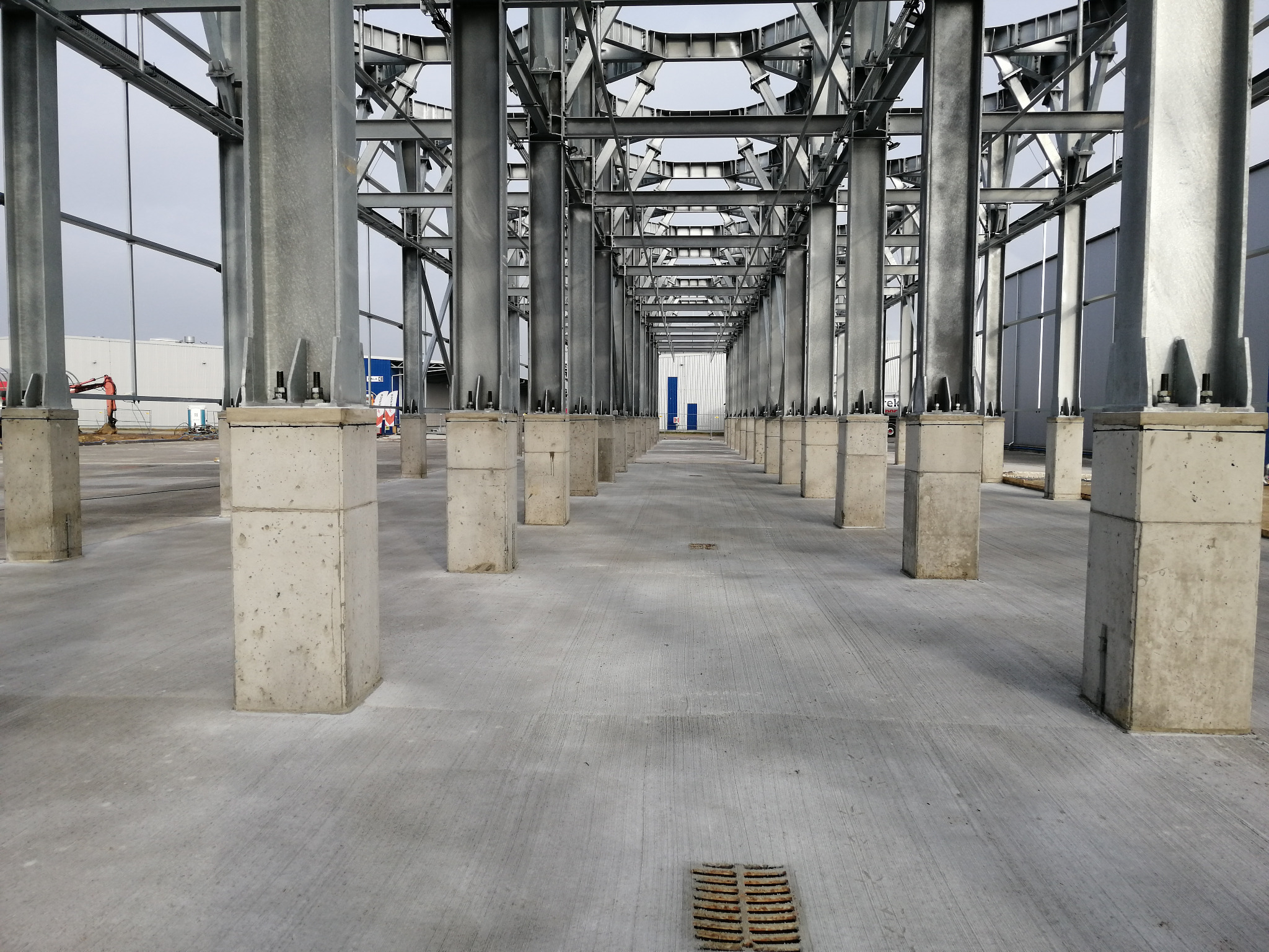 Surowa, szara posadzka przemysłowa w hali produkcyjnej z betonowymi słupami i stalową konstrukcją dachu, widoczny odpływ liniowy w podłodze.