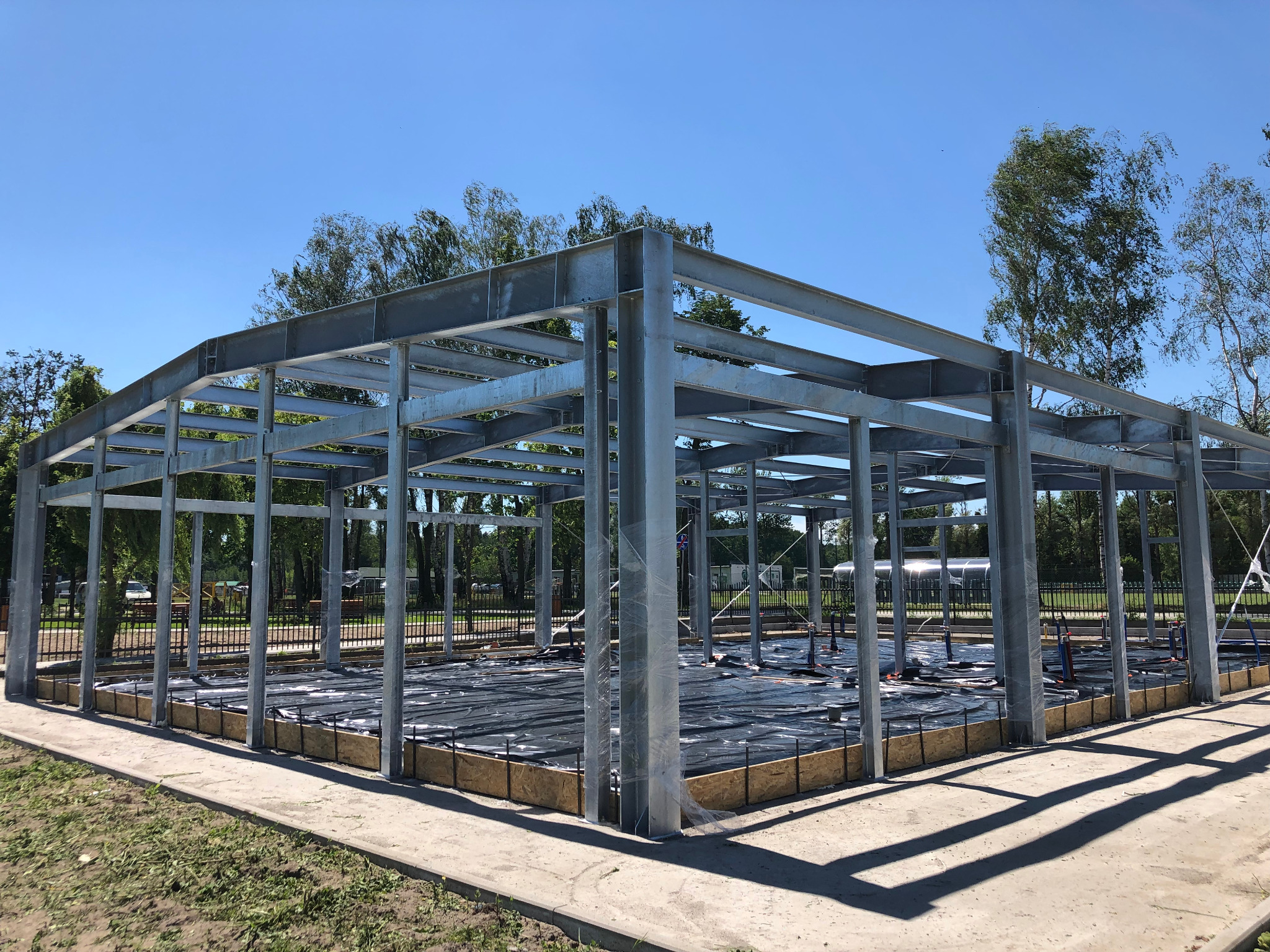 Szkielet stalowy budynku w trakcie budowy, fundament zabezpieczony czarną folią, widoczne deski szalunkowe i zbrojenie, tło z zielenią drzew i błękitnym niebem.