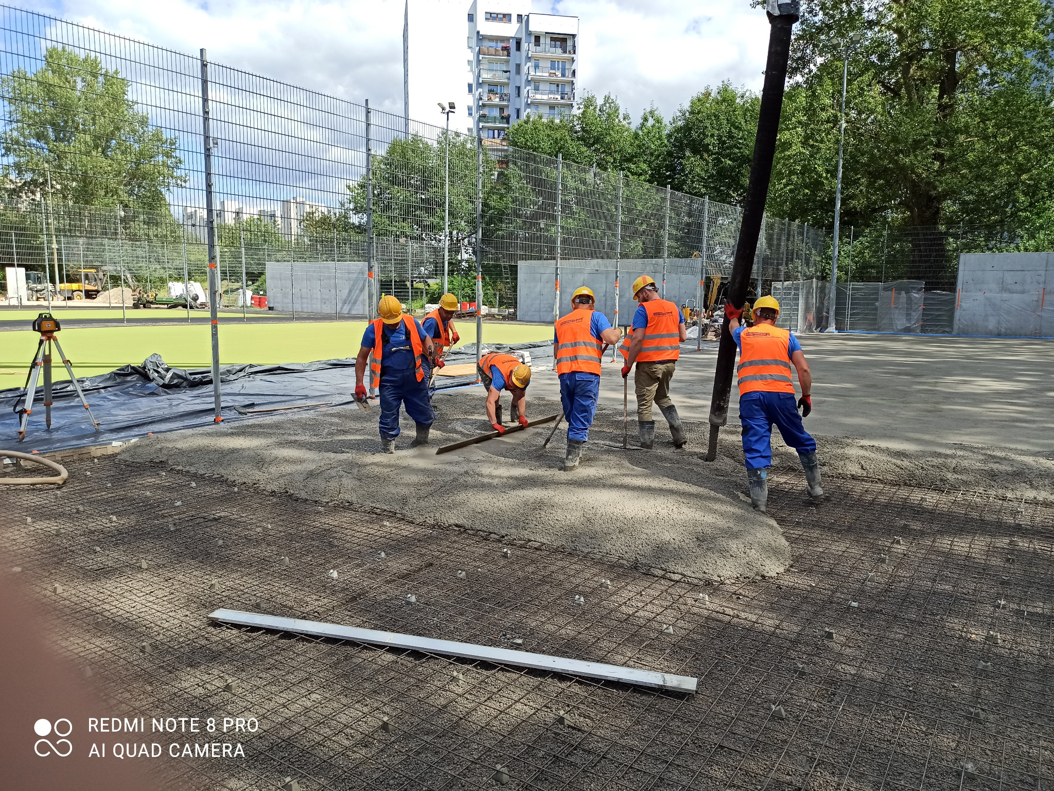 Wylewka betonowa na siatce zbrojeniowej, ekipa robotników w pomarańczowych kamizelkach rozprowadza beton, w tle boisko sportowe i budynek mieszkalny.