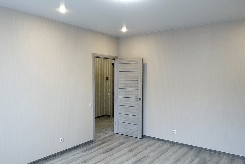 Jasne wnętrze pokoju z szarą podłogą imitującą drewno, kremowymi ścianami i otwartymi szarymi drzwiami z poziomymi panelami, widoczne gniazdka elektryczne i lampy sufitowe.