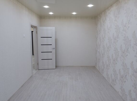 Pusty pokój po remoncie z jasnymi ścianami, wzorzystą tapetą na jednej ścianie, białą podłogą i białymi drzwiami z poziomymi, ciemnymi paskami.