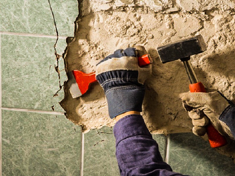 Demontaż starych, popękanych płytek ściennych w łazience przy użyciu młotka i przecinaka przez osobę w rękawicach roboczych.