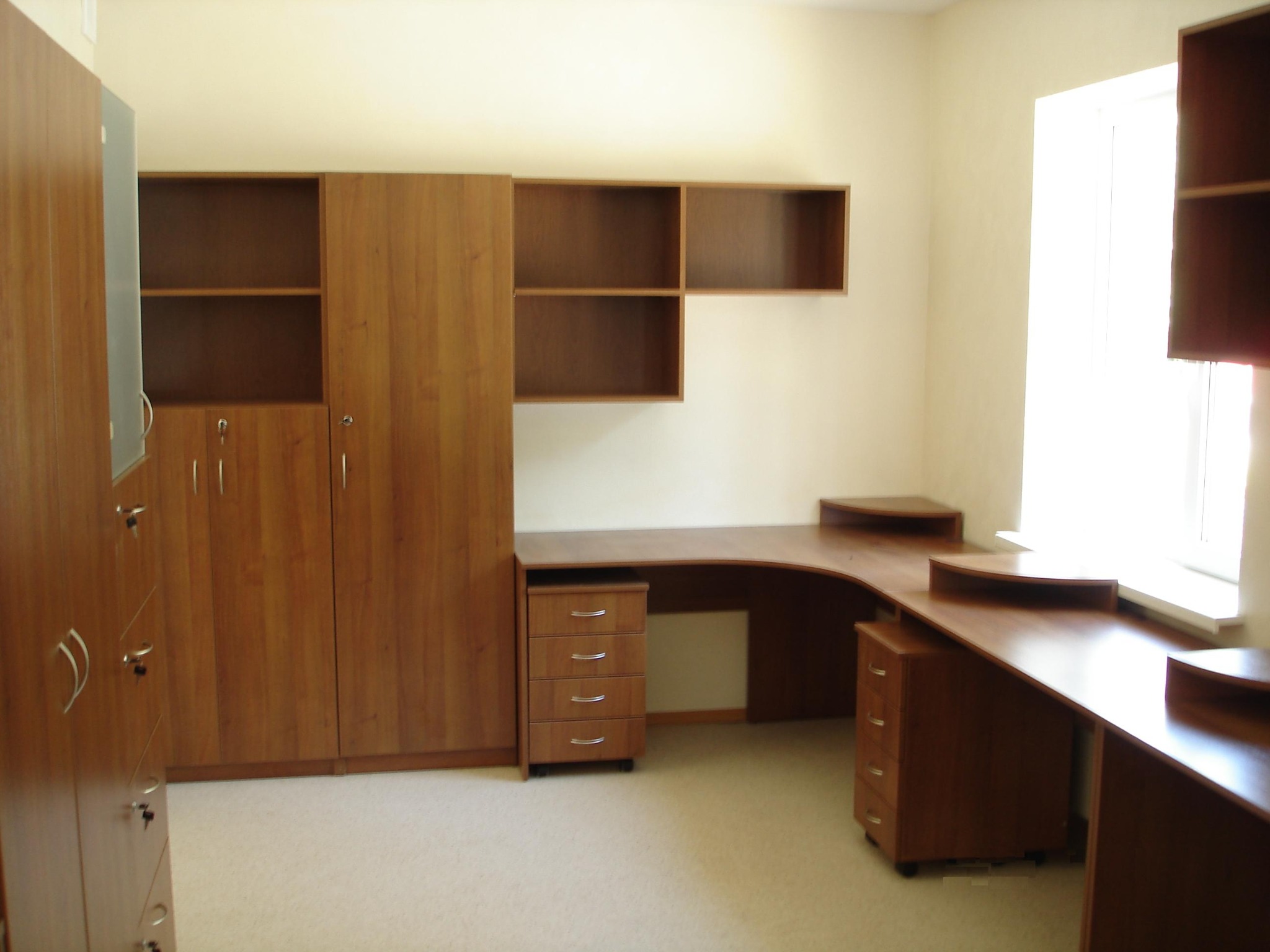 Drewniane meble biurowe w jasnym pokoju: szafki zamykane na klucz, regały i biurko narożne z szufladami, oświetlone naturalnym światłem z okna.