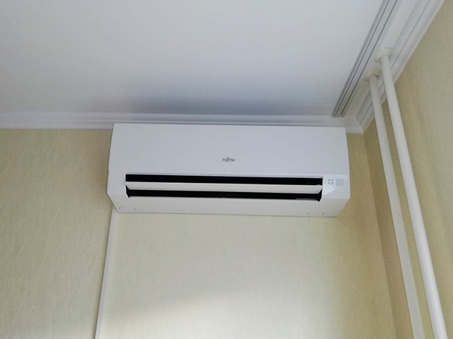 Biały klimatyzator Fujitsu zamontowany na ścianie w pomieszczeniu z kremową ścianą i białym sufitem z dekoracyjną listwą.