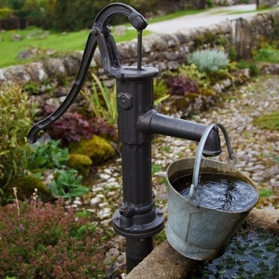 Żeliwna pompa wodna z wiadrem pełnym wody, ustawiona na kamiennej podstawie w wiejskim ogrodzie z kamiennym murem w tle.