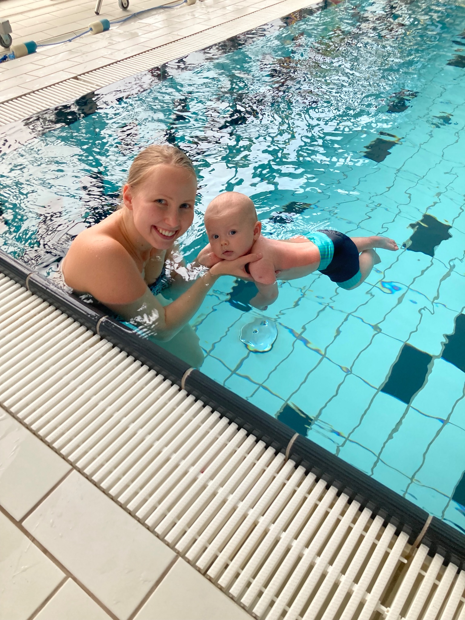 Uśmiechnięta kobieta trzymająca niemowlę w basenie z błękitną wodą i czarno-białą szachownicą na dnie.
