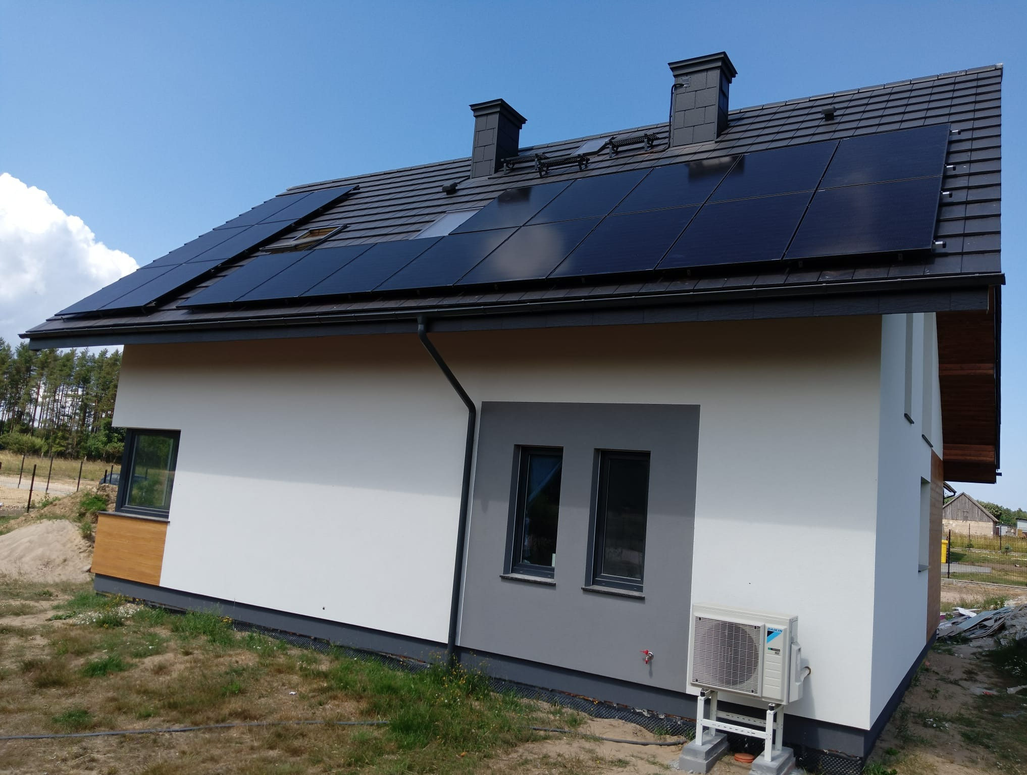 Nowoczesny dom jednorodzinny z panelami słonecznymi na dachu i jednostką zewnętrzną klimatyzacji marki Daikin, widoczne czarne kominy na dachu i szara stolarka okienna.