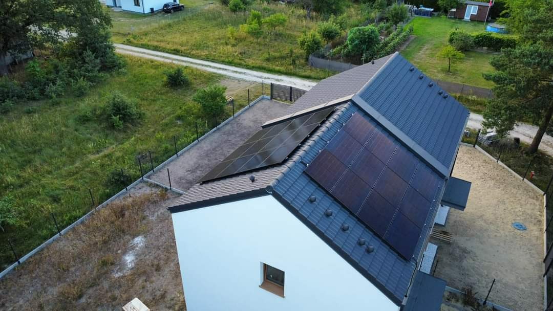 Widok z góry na dom jednorodzinny z zainstalowanymi panelami fotowoltaicznymi na dachu, otoczony zielenią i ogrodzeniem z siatki.