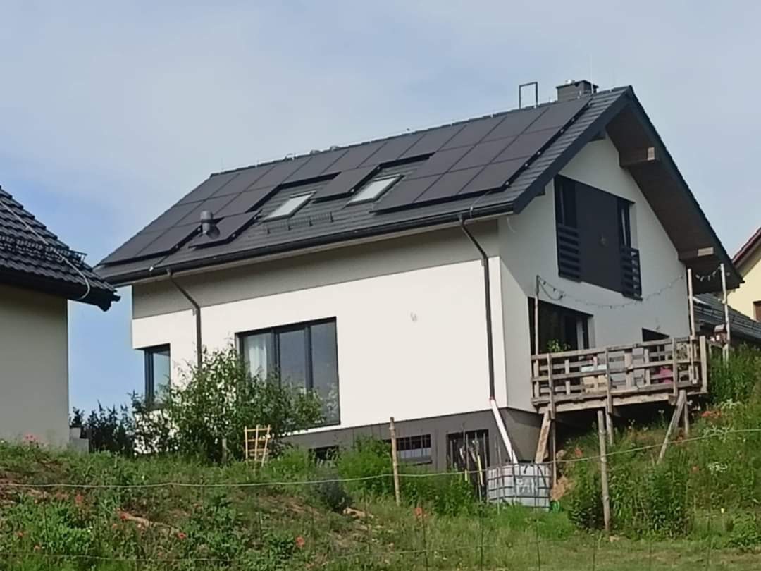 Dom jednorodzinny z panelami słonecznymi na dachu, widoczne okna dachowe i rynna odprowadzająca wodę do zbiornika na deszczówkę.