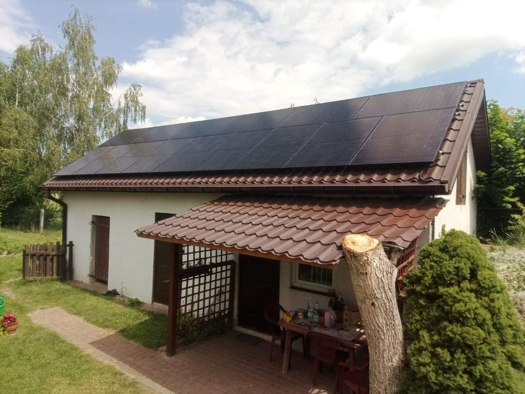 Dom jednorodzinny z brązowym dachem i panelami słonecznymi na dachu, widok z poziomu trawnika, obok pnia ściętego drzewa.