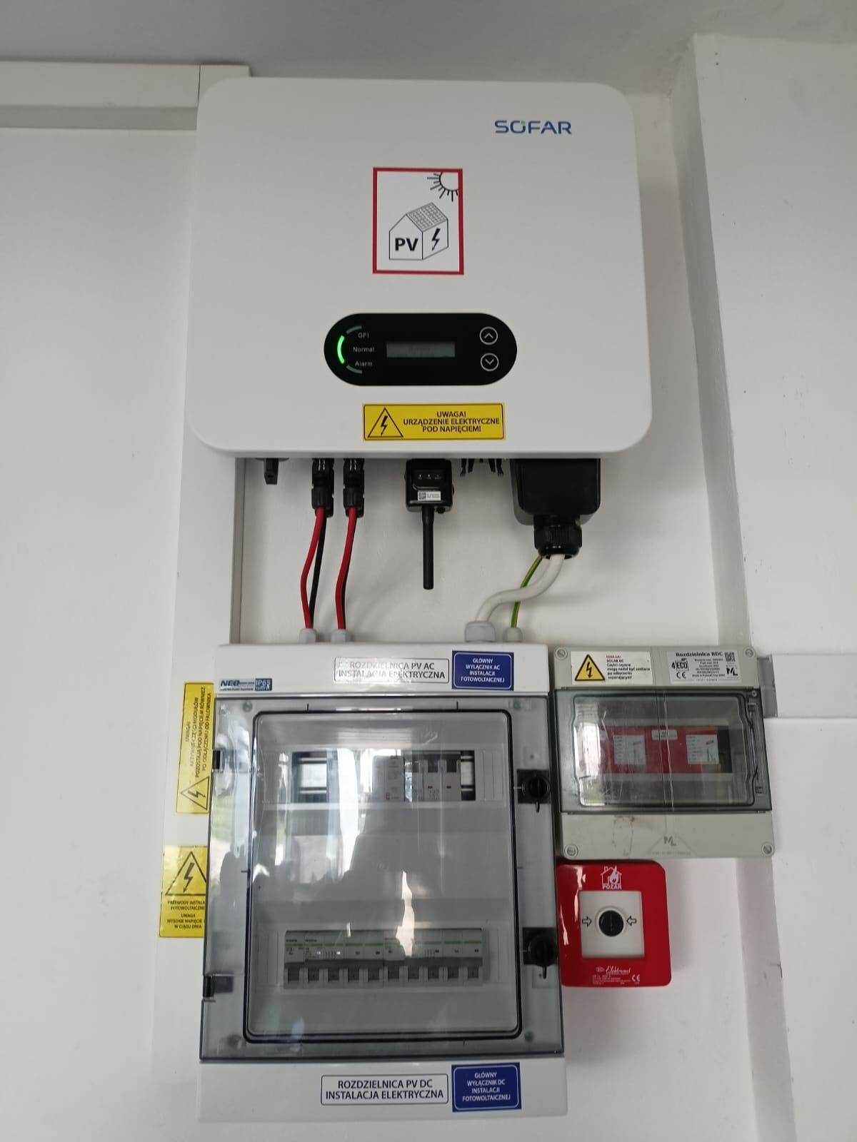 Biały falownik SOFAR z podłączonymi przewodami i rozdzielnicami PV AC i DC na białej ścianie, widoczne ostrzeżenia o napięciu.