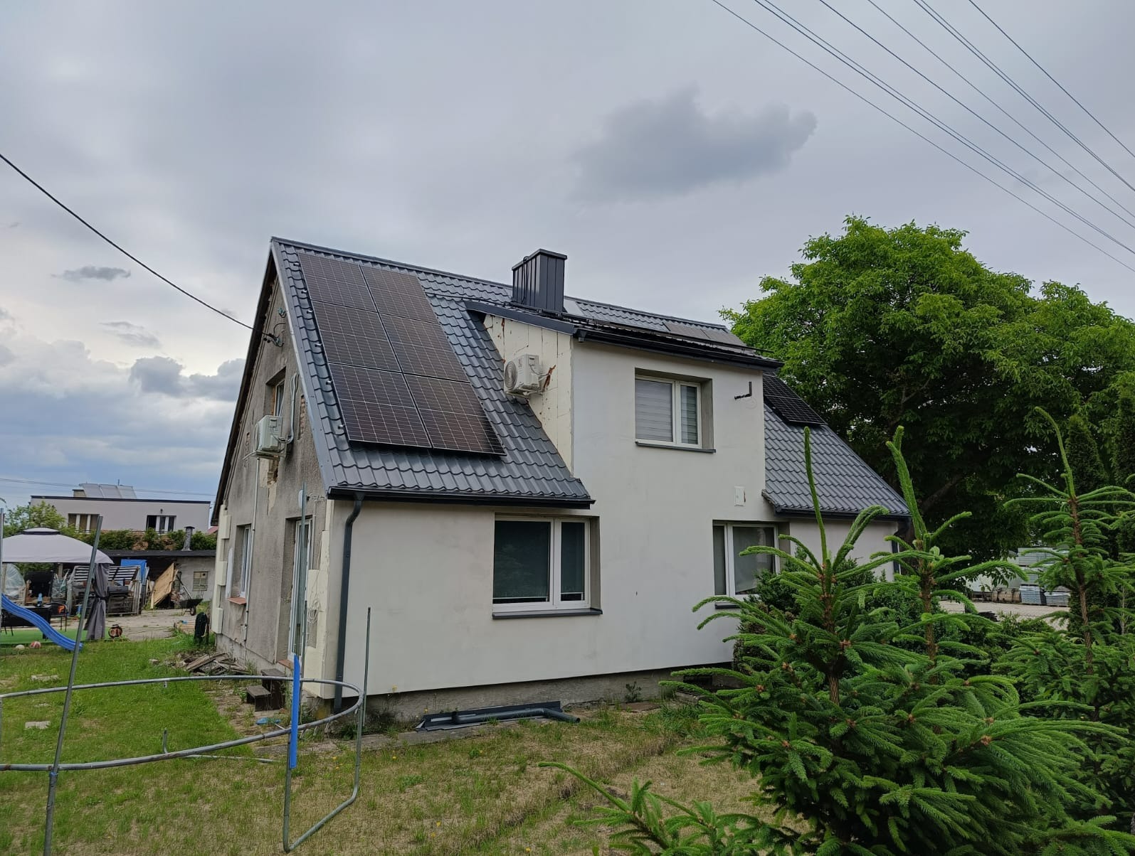 Dwukondygnacyjny dom z panelami słonecznymi na dachu z szarej dachówki, widoczne również dwa klimatyzatory na elewacji, pochmurne niebo w tle.