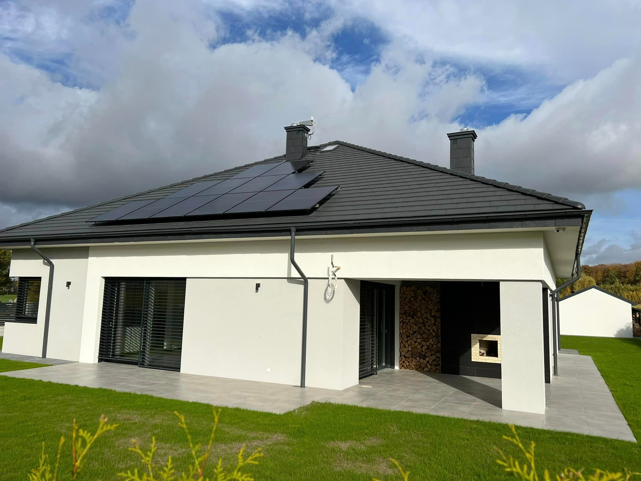 Nowoczesny dom jednorodzinny z panelami słonecznymi na grafitowym dachu i zapasem drewna kominkowego pod zadaszeniem.