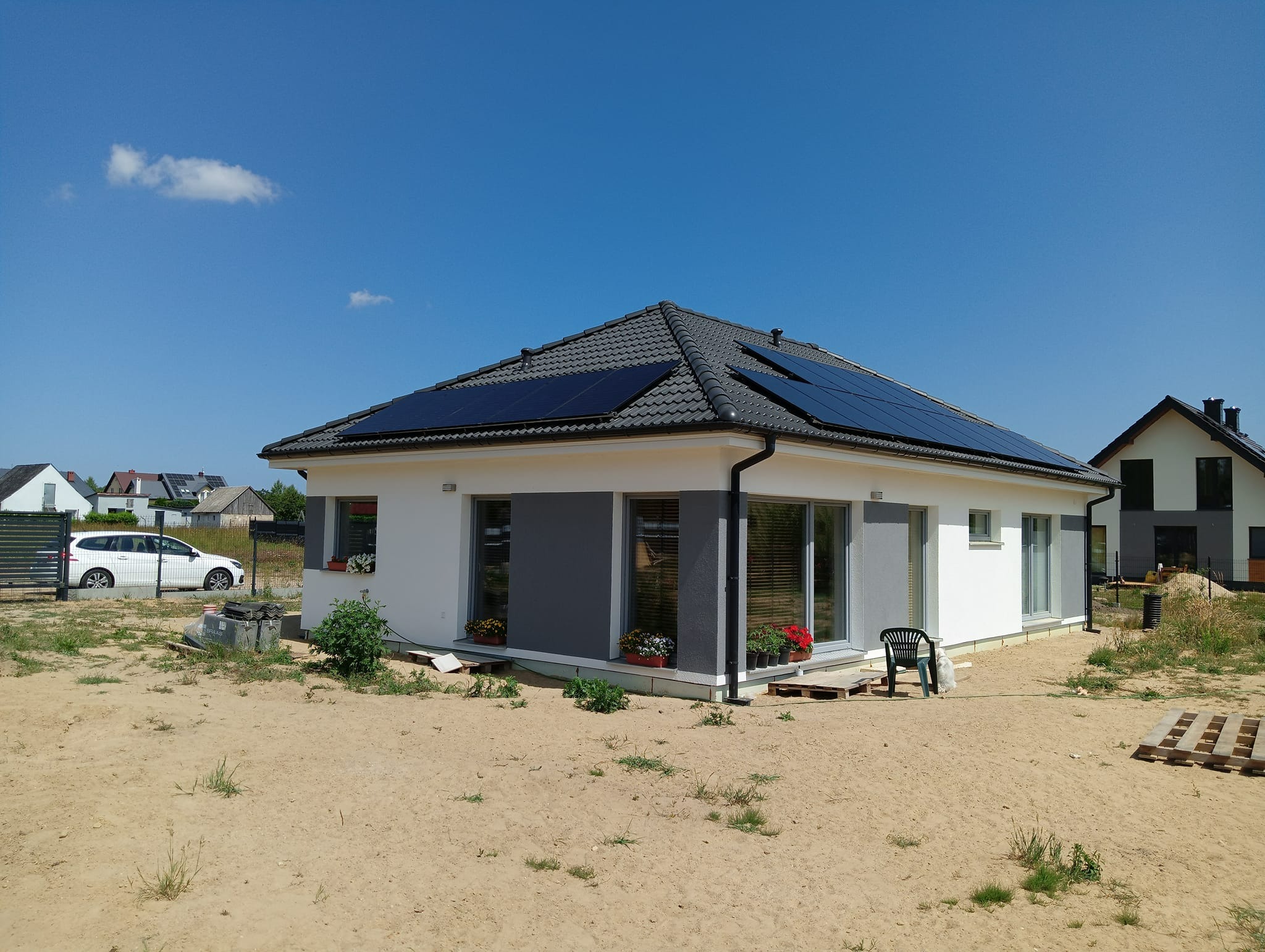 Nowoczesny dom jednorodzinny z panelami słonecznymi na dachu, w tle błękitne niebo, teren wokół domu w trakcie zagospodarowywania.