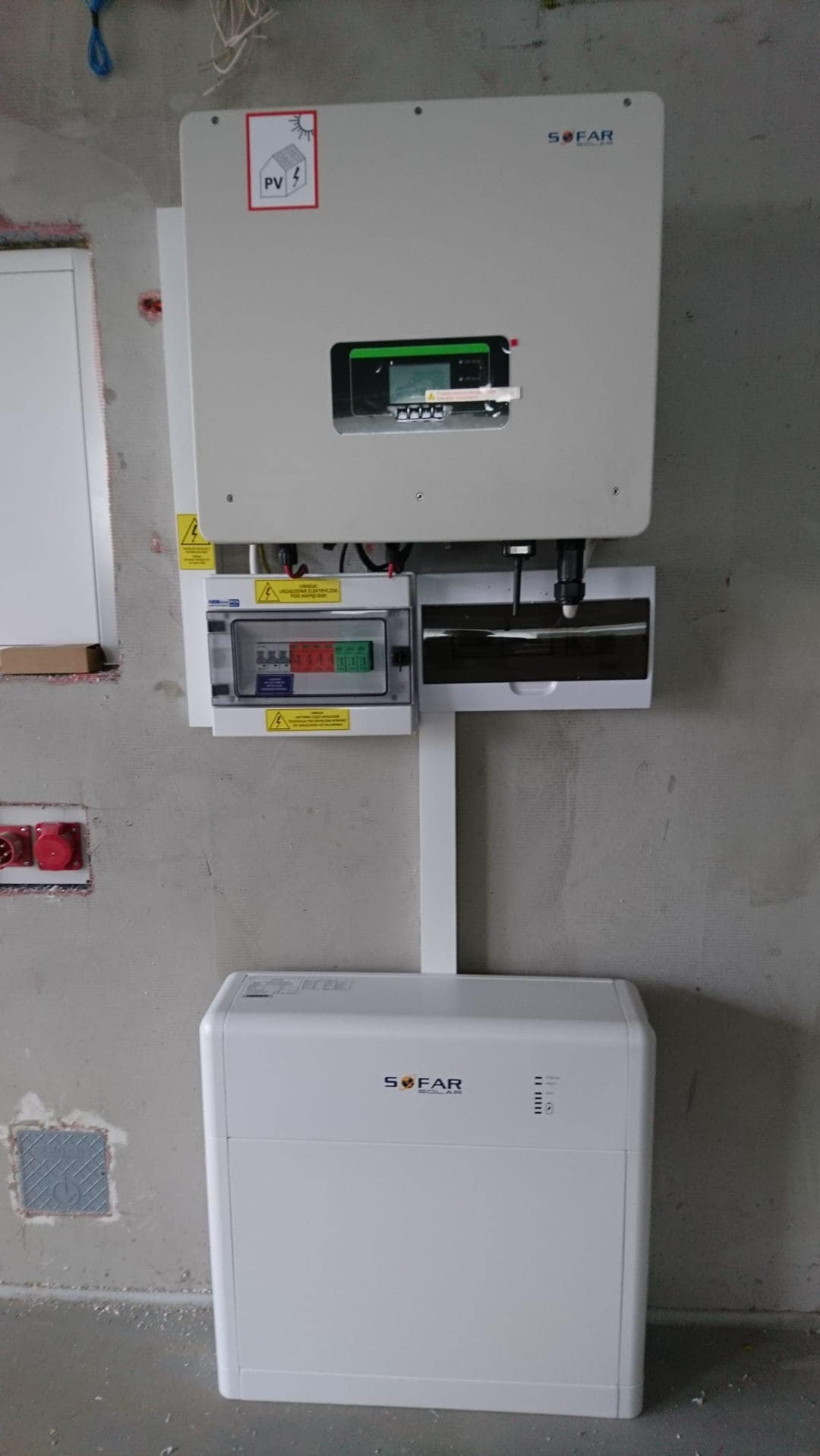 Zamontowany biały falownik z wyświetlaczem i rozdzielnica elektryczna na surowej ścianie, obok widoczne gniazda elektryczne.