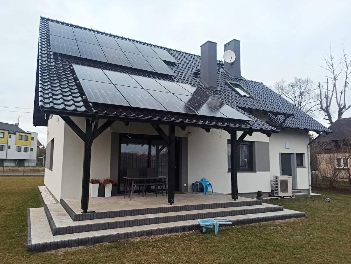 Dom jednorodzinny z panelami fotowoltaicznymi na dachu, widoczny z perspektywy ogrodu, z zadaszonym tarasem i pompą ciepła na zewnątrz budynku.