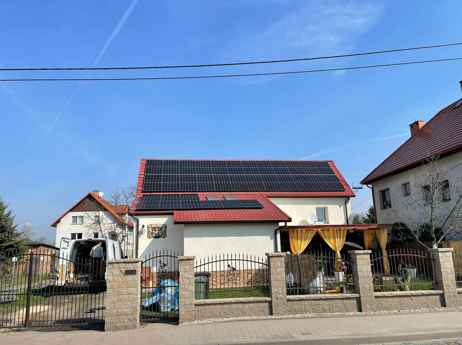 Dom jednorodzinny z panelami słonecznymi na czerwonym dachu; w tle błękitne niebo z widocznymi smugami kondensacyjnymi; z otwartego bagażnika białego samochodu dostawczego wyjmuje sprzęt instalator.