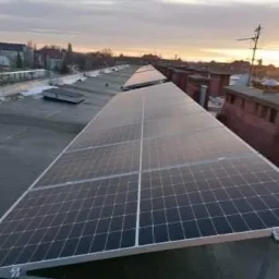 Rząd paneli słonecznych na płaskim dachu budynku w pochmurny dzień, widoczny zachód słońca na horyzoncie.