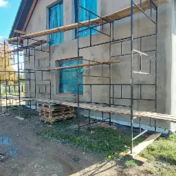 Surowa elewacja domu jednorodzinnego w trakcie tynkowania, okna zabezpieczone folią, rusztowanie metalowe z drewnianymi pomostami, palety budowlane obok rusztowania.