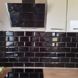 Czarne, błyszczące płytki ułożone na ścianie w kuchni, z widocznym okapem Gorenje i fragmentem blatu z drobnymi opiłkami.