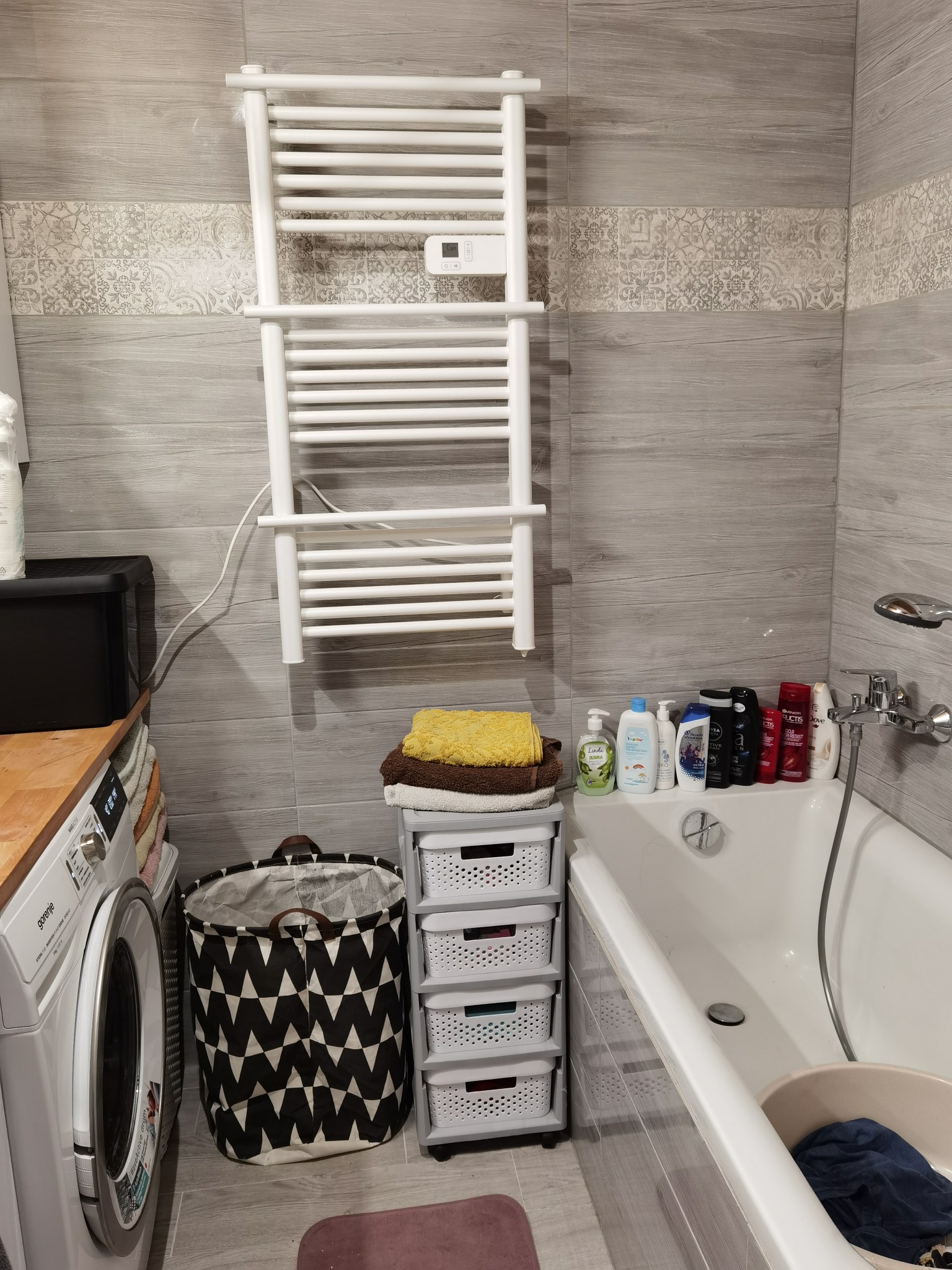 Uporządkowana łazienka z pralką, koszem na pranie, ręcznikami, grzejnikiem drabinkowym i kosmetykami na wannie.