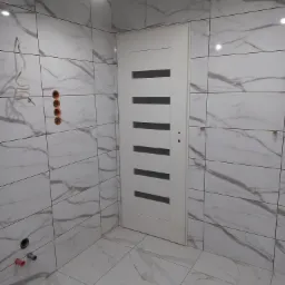 Łazienka w trakcie remontu: białe płytki imitujące marmur na ścianach i podłodze, białe drzwi z poziomymi przeszkleniami, widoczne instalacje elektryczne i hydrauliczne przed wykończeniem.