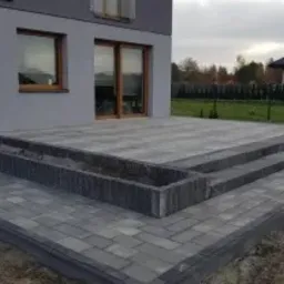 Szara kostka brukowa ułożona na tarasie przed nowoczesnym domem z szarymi ścianami i dużymi oknami tarasowymi. Widoczne trzy stopnie schodów i obrzeże rabaty kwiatowej, również wyłożone kostką.