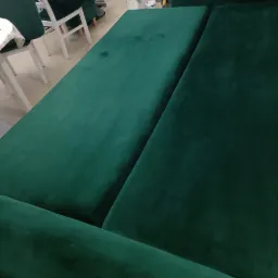 Szmaragdowa, welurowa kanapa po praniu, widoczne delikatne smugi wilgoci w górnym rogu siedziska, jasne krzesła i odkurzacz w tle.