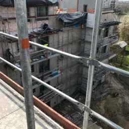 Budynek wielorodzinny w trakcie prac elewacyjnych, widok z rusztowania, pracownicy montują izolację, zabezpieczenie folią na górnych kondygnacjach, wiosenne drzewa w tle.