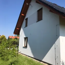 Świeżo otynkowany dom jednorodzinny z białą elewacją, brązową stolarką okienną i ciemną dachówką, widok z ogrodu w słoneczny dzień.